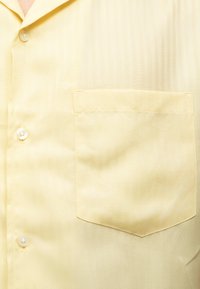 Ανοιχτό κίτρινο πουκάμισο με γιακά, τσέπη στο στήθος και τρία λευκά κουμπιά στην μπροστινή πατιλέτα.