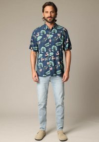 Korte mouw navy shirt met een tropische bladeren- en bloemenprint; gecombineerd met lichtblauwe jeans en beige instappers.