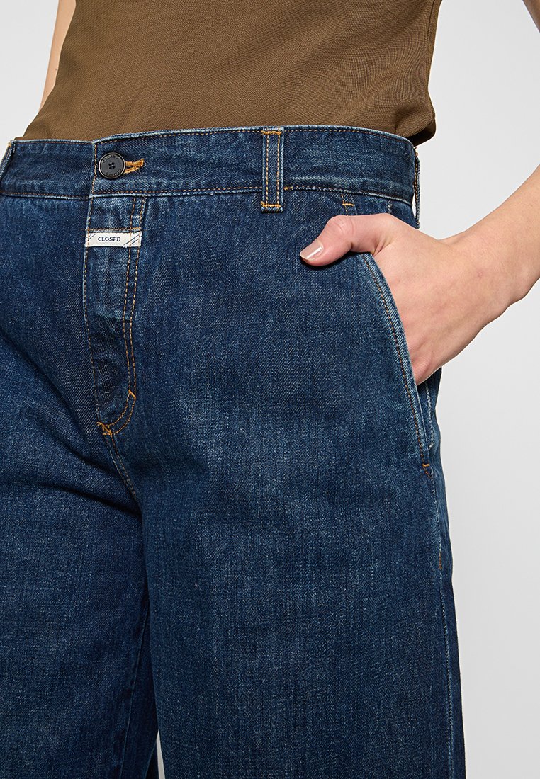 Jeans dżinsowe w ciemnoniebieskim kolorze z żółtymi przeszyciami, przednią kieszenią oraz pasem z zapięciem na guzik i metką marki.