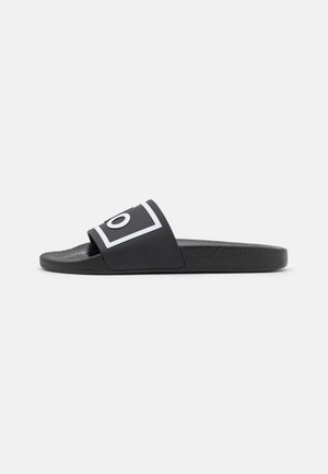 Schwarze Slidesandale mit strukturiertem Sohlenprofil und einem breiten Riemen, der ein weiß umrandetes Quadrat mit dem Buchstaben "O" zeigt.
