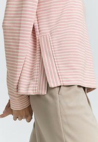 Maglietta a maniche lunghe a righe rosa e bianche con fessure laterali, realizzata in tessuto morbido. Abbinata a pantaloni beige, che mostrano un design casual.