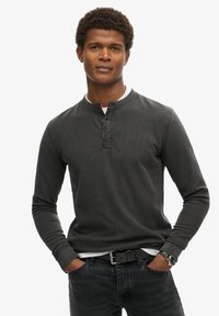 Camisa henley de manga larga gris oscuro con una tela texturizada, tapeta de tres botones y cuello redondo, combinada con jeans oscuros y un cinturón.