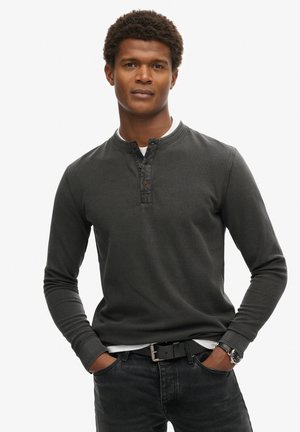 Camisa henley de manga larga gris oscuro con una tela texturizada, tapeta de tres botones y cuello redondo, combinada con jeans oscuros y un cinturón.