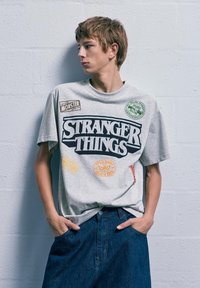 Szary bawełniany T-shirt z grafiką "STRANGER THINGS" w kolorze czarnym, z kolorowymi naszywkami i logo. W połączeniu z ciemnymi szortami dżinsowymi.