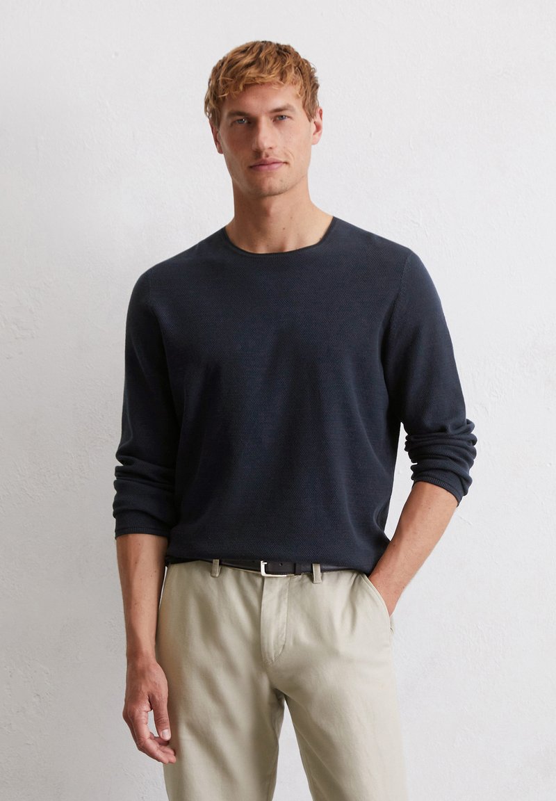 Marineblauer Strickpullover mit langen Ärmeln und Rundhalsausschnitt, kombiniert mit hellbeigen Hosen und einem schwarzen Gürtel. Weiche Textur, mittlere Passform und hochgerollte Ärmel.