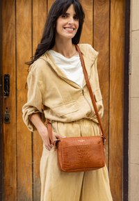 Brązowa torba crossbody w wzór krokodyla z teksturowanym wykończeniem i długim paskiem. Noszona z beżowym ubraniem na tle drewnianym.