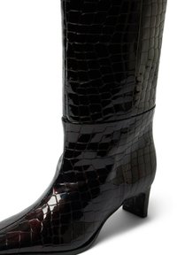 Põlveni ulatuvad mustad patendi-nahast saapad, millel on läikiv krokodillinahk, terav nina ja madal ristkülikukujuline konts.