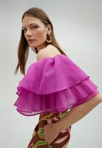Off-shoulder top med lagdelte volanger i levende fuchsia. Brun mønstrede stof med grønne og lyserøde accenter. Model holder en lomme.