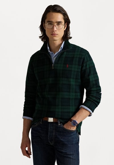 Grønternet zip-hoodie lavet af blødt stof, lagdelt over en lyseblå skjorte med krave. Mørke jeans og et sølvbrunt ur fuldender looket.