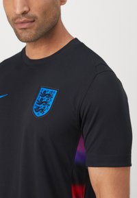 Juodos trumpomis rankovėmis sportinė marškinė. Krūtinėje yra mėlynas Anglijos heraldikos ženklas, o šone matomas violetinis gradientinis akcentas.