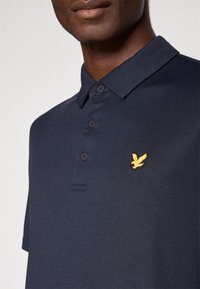 Tamsiai mėlynas polo marškinėlis, pagamintas iš tekstūruoto audinio, su trimis mygtukais priekyje ir auksiniu erelio logotipu kairiajame krūtinės šone.