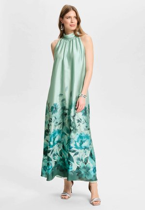 Femme portant une robe longue sans manches en satin vert menthe avec un motif floral près de l'ourlet, des bracelets argentés et des sandales à talons hauts.