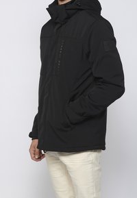 Veste noire isolante avec capuche, dotée de poches zippées, de panneaux texturés et d'un logo à l'avant. Fabriquée en matériau durable.