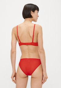 Piros fehérneműszett áttetsző anyaggal, állítható pántokkal és csipkés szegéllyel a bugyiban. Hátulnézet a minimalista dizájnt emeli ki.