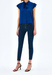 Blauwe blouse met korte mouwen, een kraag en strikdetail, gecombineerd met donkere skinny jeans en zwarte sandaaltjes met banden. Gladde textuur, aansluitend ontwerp.