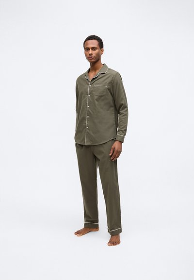 Man in een olijfgroen pyjama met lange mouwen en knoopsluiting, gecombineerd met bijpassende broek met witte biezen, staand op blote voeten tegen een plain witte achtergrond.