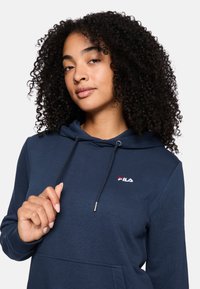 Sudadera color azul marino con capucha ajustable, puños acanalados y un bolsillo frontal. Presenta un logo bordado en rojo y blanco en el pecho.