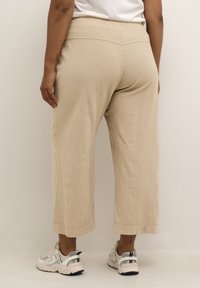 Pantalons courts de couleur beige, légers, avec une texture lisse, une taille élastique et des détails de couture latérale, associés à des baskets claires.