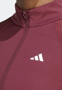 Närbild av en bourgognaröd zip-jacka med hög krage och en liten vit geometrisk logotyp på bröstet.