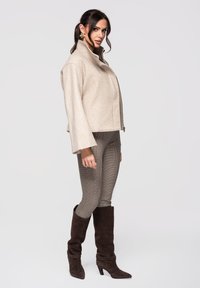 Femme portant une courte veste beige, un pantalon slim à motifs, des bottes marron foncé à talons montant jusqu'aux genoux et des boucles d'oreilles dorées, debout devant un fond clair.