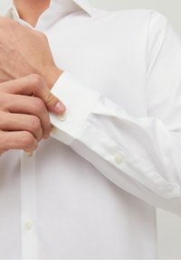 Camicia bianca con una texture liscia, polsini con bottone e colletto classico. Il tessuto appare leggero e traspirante.