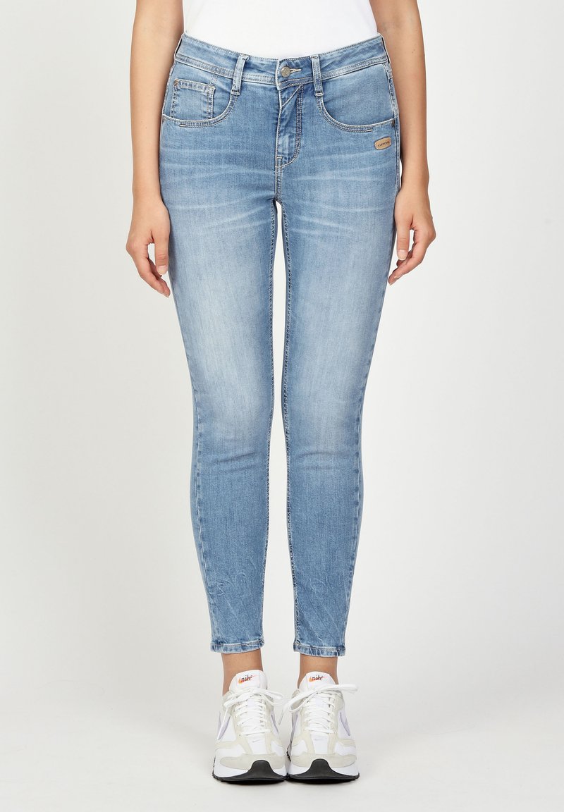 Light blue skinny jeans made of denim, featuring a mid-rise waist, five-pocket design, and subtle fading. White sneakers visible at the bottom.