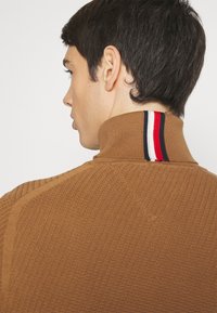 Pullover marrone con un motivo a maglia testurizzata, colletto alto e una striscia tricolore in rosso, bianco e blu sulla parte posteriore.