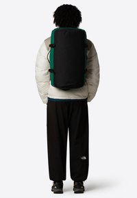 Sac à dos noir et vert de forme cylindrique, avec des bretelles réglables, un tissu robuste et des détails matériels minimaux, porté sur une veste blanche volumineuse.