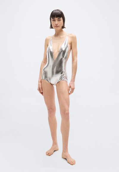 Γυναίκα που στέκεται ξυπόλητη, φορώντας μια ολόσωμη μεταλλική ασημένια μαγιό με βαθιά V λαιμόκοψη, μπροστά σε απλό φωτεινό φόντο.