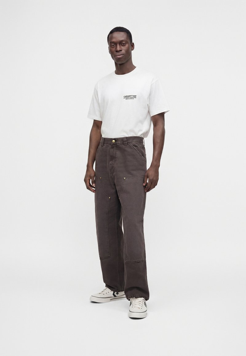 Mann steht, trägt weißes T-Shirt mit kurzen Ärmeln und Text sowie dunkelbraune, lockere Hose mit aufgesetzten Taschen und weiße Sneakers vor weißem Hintergrund.