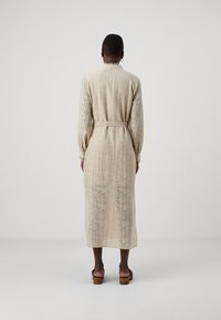 Long, beige, textured dress with a subtle leaf pattern, belted at the waist, featuring long sleeves and a collar. Worn with wooden-heeled sandals.