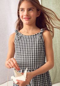 Ragazza che indossa un vestito a quadretti bianco e nero con spalline annodate, che tiene un origami di carta a forma di gru su uno sfondo morbido e neutro.