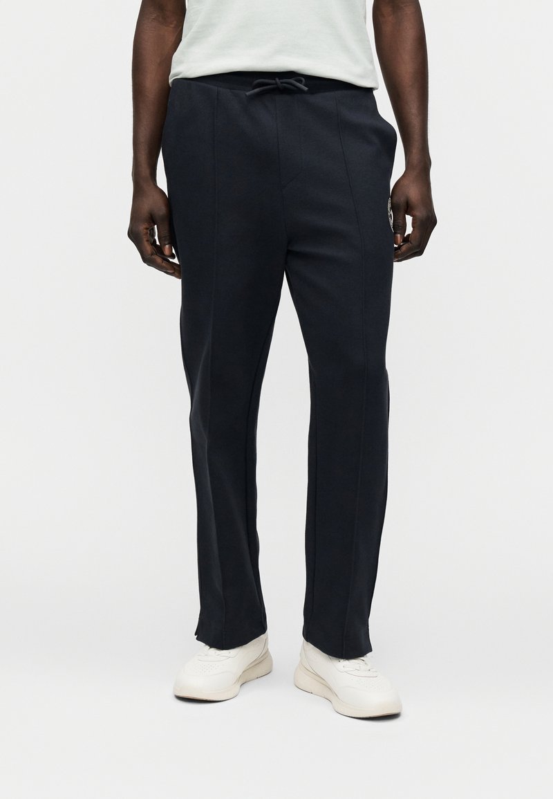 Σκούρες μπλε φόρμες από μαλακό ύφασμα, με κορδόνι στη μέση, πλευρικές ραφές και λογότυπο στην αριστερή μηρό.