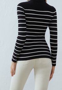 Jersey de cuello alto a rayas en blanco y negro, textura acanalada, diseño ajustado, combinado con pantalones ajustados de color claro. Fondo de color sólido.