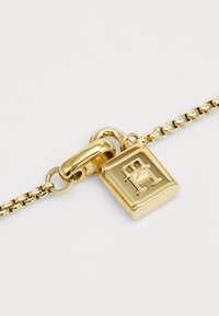 Pulsera de cadena dorada con un diseño de candado y un logotipo grabado. La cadena está formada por eslabones ovalados entrelazados, creando una apariencia texturizada.