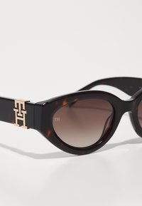 Brown tortoiseshell sunglasses with gradient lenses, featuring a gold-tone "TH" logo on the frame and a cat-eye design.