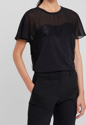 Vrouw draagt een zwart doorschijnend kortmouwig shirt met kantdetails, gecombineerd met zwarte high waist broeken, hand in de zak.
