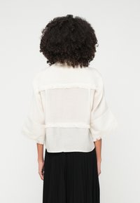 White kimono-style top with wide sleeves, featuring a frayed hem and sheer panel at the back. Worn over a black skirt.