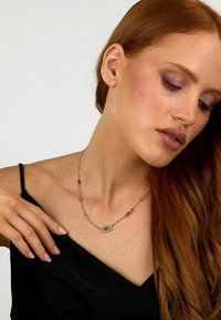 Collana color oro con pietra centrale rettangolare, circondata da collegamenti decorativi; indossata su una spalla scoperta; finitura liscia e lucida.