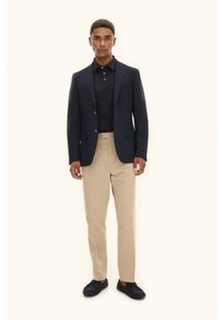 Marinblå kavaj med två knappar, mörk pikétröja, beige byxor och mörka loafers; slät tyg, skräddarsydd passform och klassisk design.