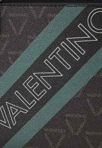 Matériau noir texturé avec une bande diagonale verte, présentant le logo "VALENTINO" en blanc. Des motifs du nom de marque répété sont visibles en arrière-plan.