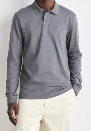 Homme portant un polo gris à manches longues avec un logo sur la poitrine et un pantalon beige clair, debout devant un fond uni.