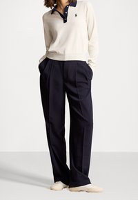 Polo beige à manches longues avec col foncé, associé à un pantalon plissé bleu marine et des sneakers clairs. Tissu doux, coupe décontractée, design casual.