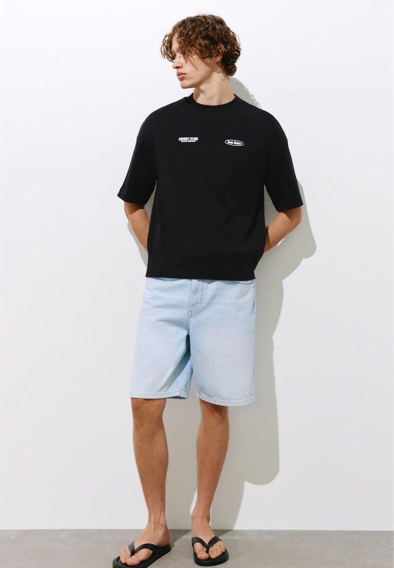 Jeune homme aux cheveux bouclés portant un t-shirt noir à motif, un short en jean clair et des tongs noires, debout contre un mur blanc.