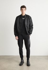 Crna bomber jakna, crna majica kratkih rukava, crne sportske hlače s logom i crne gležnjače. Glatka tekstura, ležeran kroj i čist dizajn.
