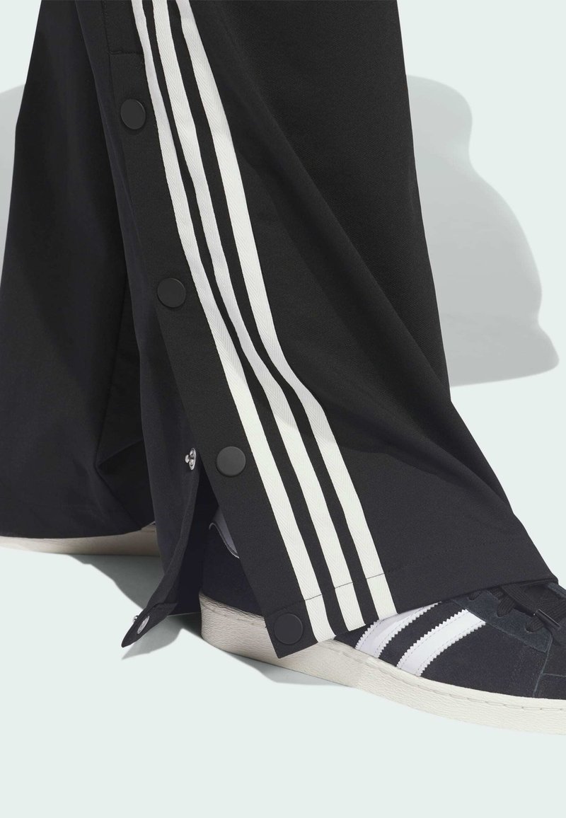 Black athletic pants with white stripes, snap buttons on the side, paired with black sneakers featuring white accents.
