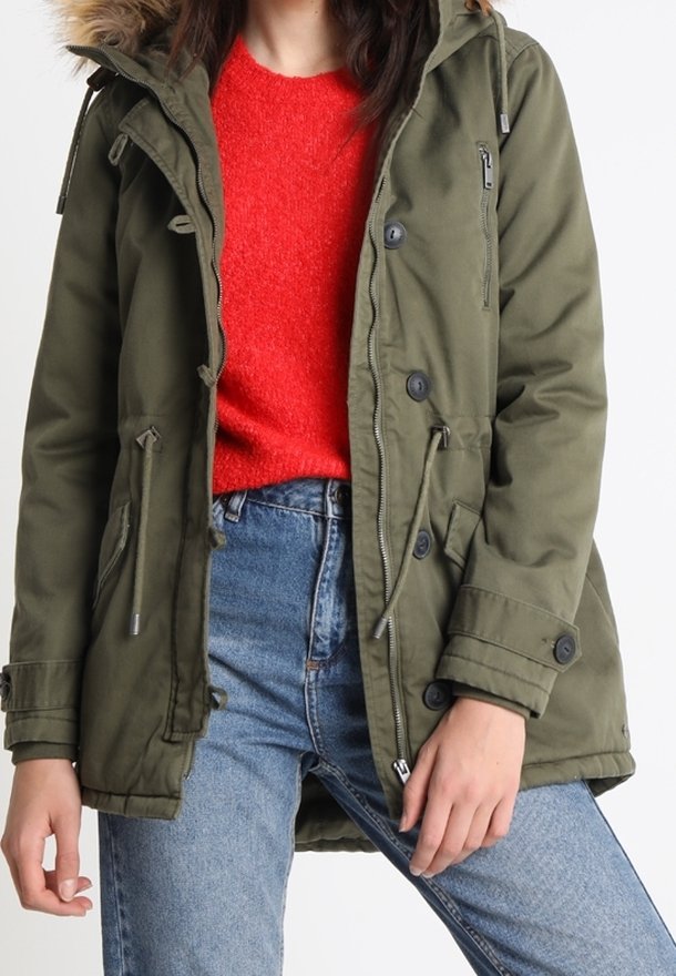 Grön parka-jacka med huva i syntetisk päls, med knappar, dragsko och flera fickor. Bärs över en röd tröja och blå jeans.