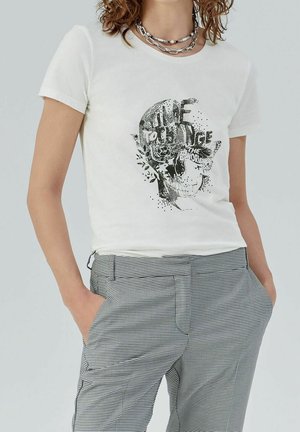 Femme portant un t-shirt blanc avec un motif crâne et papillon et le texte « Time to change the world », associé à un pantalon à carreaux noirs et blancs.