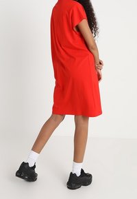 Robe rouge à manches courtes, avec une coupe ample, associée à des baskets noires et des chaussettes blanches, se tenant sur un fond uni.