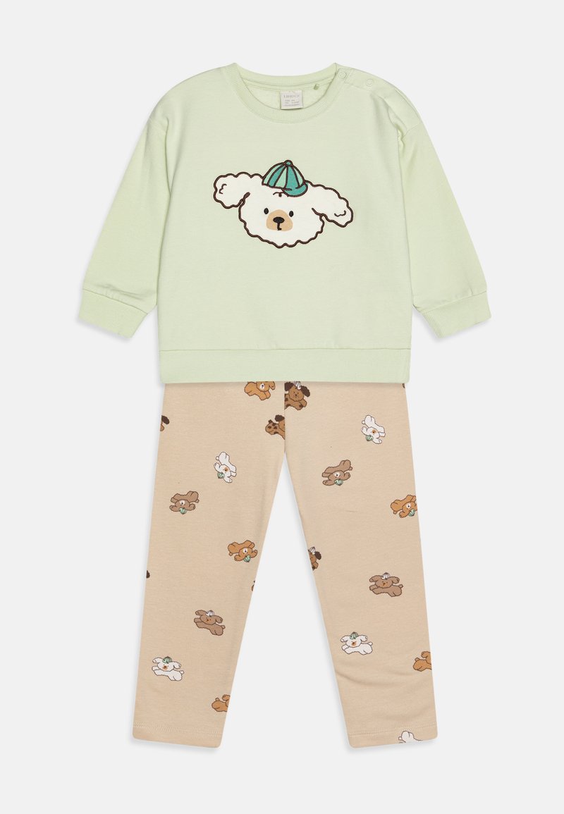 Sweatshirt vert clair avec un motif de chien et une casquette bleue, associé à un pantalon beige orné de divers motifs de chiens. Matière douce en coton.